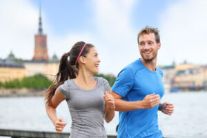 Smiling man and woman jogging outdoors near water with city buildings in the background.