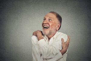 Smiling elderly man hugging himself while looking upward, wearing a white shirt against a gray background.