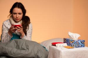 Sick woman sitting in bed with a scarf around her neck, holding a cup. A tissue box, pills, and a plate are on the bedside table.