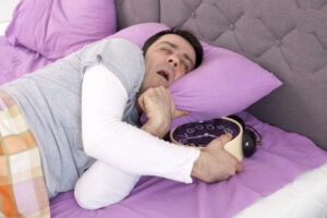 Man sleeping in bed while hugging a large alarm clock, eyes closed and mouth open.
